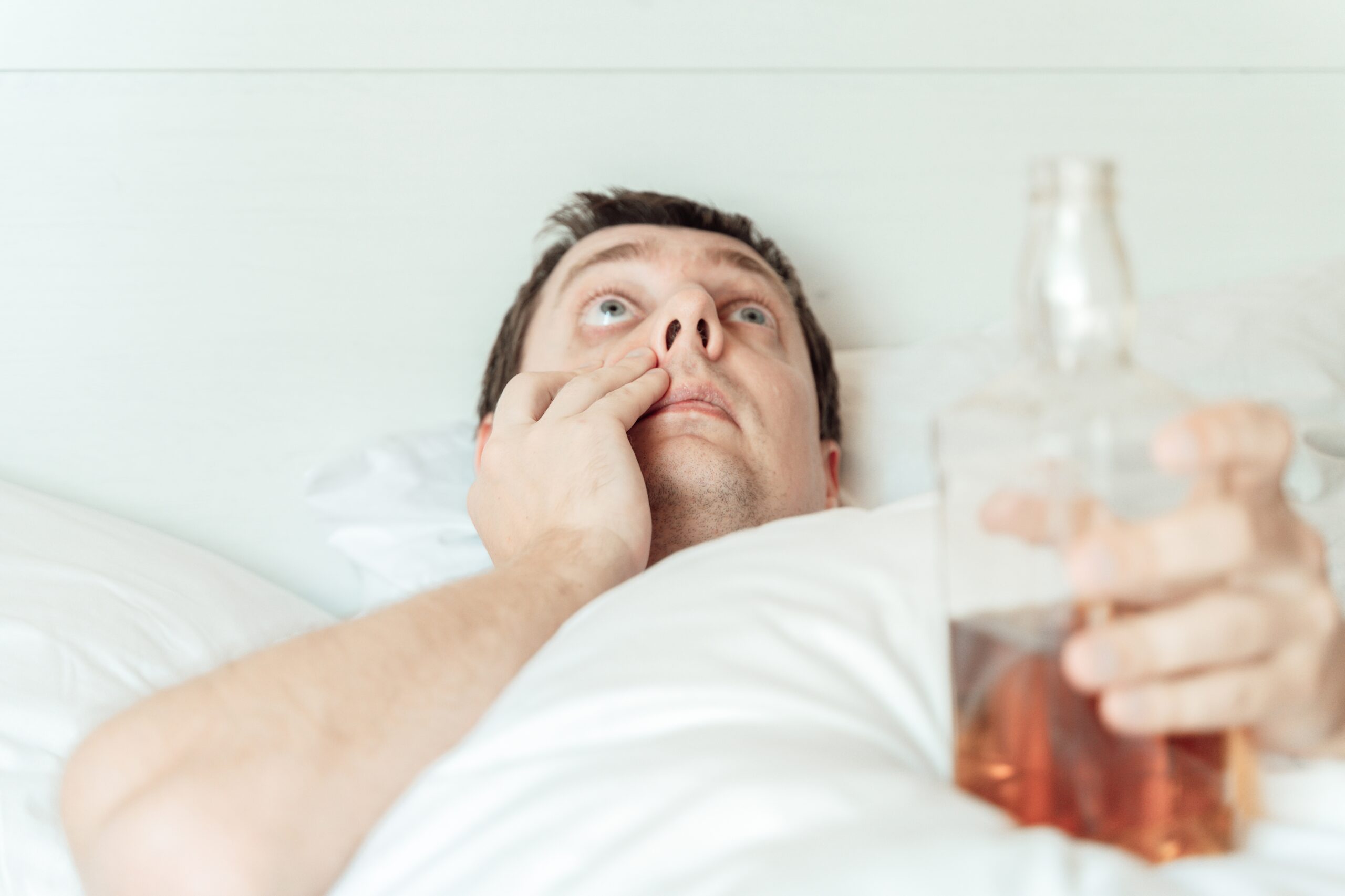 a man in bed with an alcohol bottle and one hand pulling his eyelid down, highlighting the battle between alcohol vs sleep