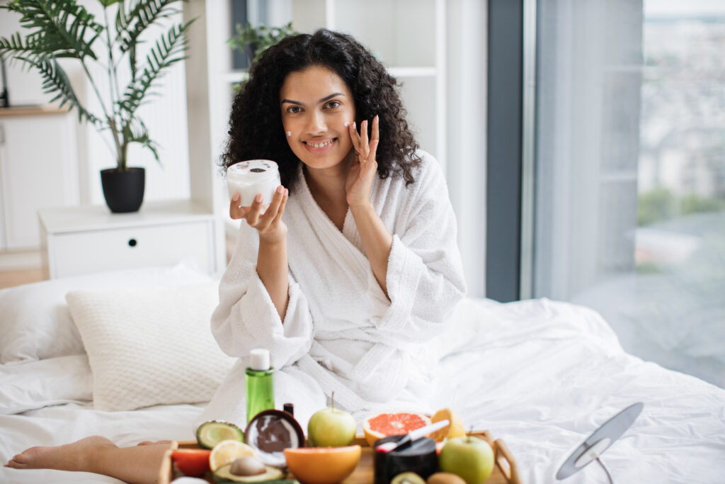 a woman sitting on her bed in a robe surrounded by health foods and creams for her face, symbolizing the importance of National Self Care Day