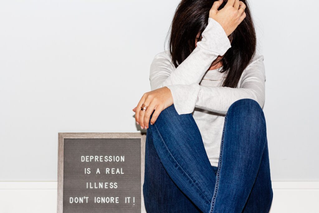 a woman sitting with her hand on her head next to a sign that says "depression is a real illness, don't ignore it", highlighting the need for depression and addiction treatment